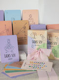 A yoga card deck set with multiple pastel-colored cards featuring minimalist line drawings of yoga poses and Hebrew text, arranged on a light surface with a pink ribbon bow and a small white box.