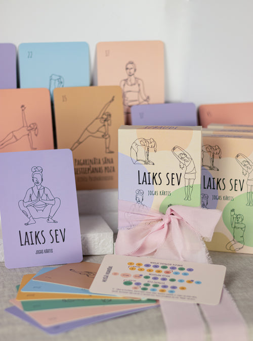 A yoga card deck set with multiple pastel-colored cards featuring minimalist line drawings of yoga poses and Hebrew text, arranged on a light surface with a pink ribbon bow and a small white box.