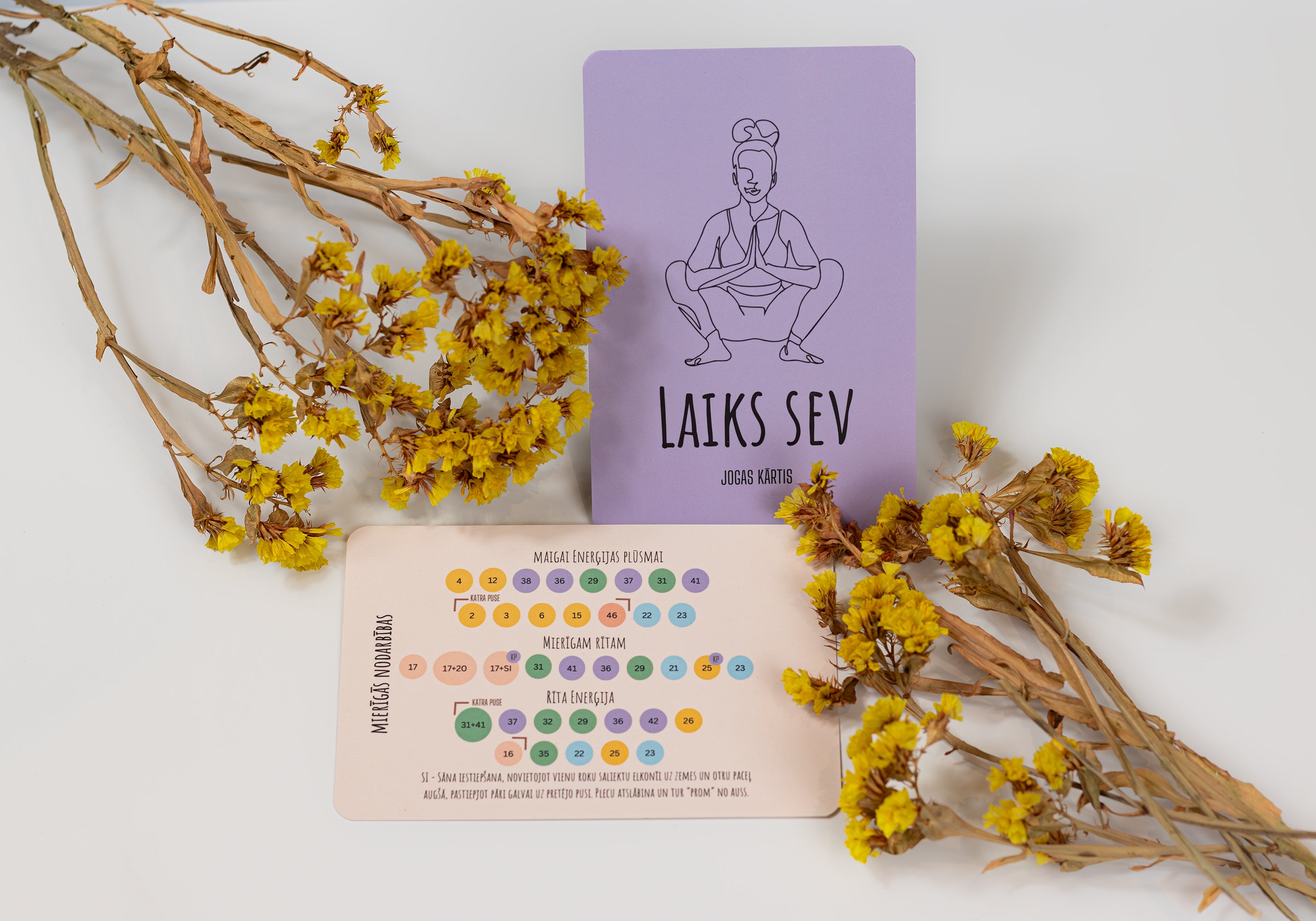 A flat-lay product shot of a 'Laiks Sev' yoga card deck by Laiks Sev featuring a lavender card with a black line drawing of a yoga pose and a beige card with colorful circular icons, styled with dried yellow flowers on a white background.