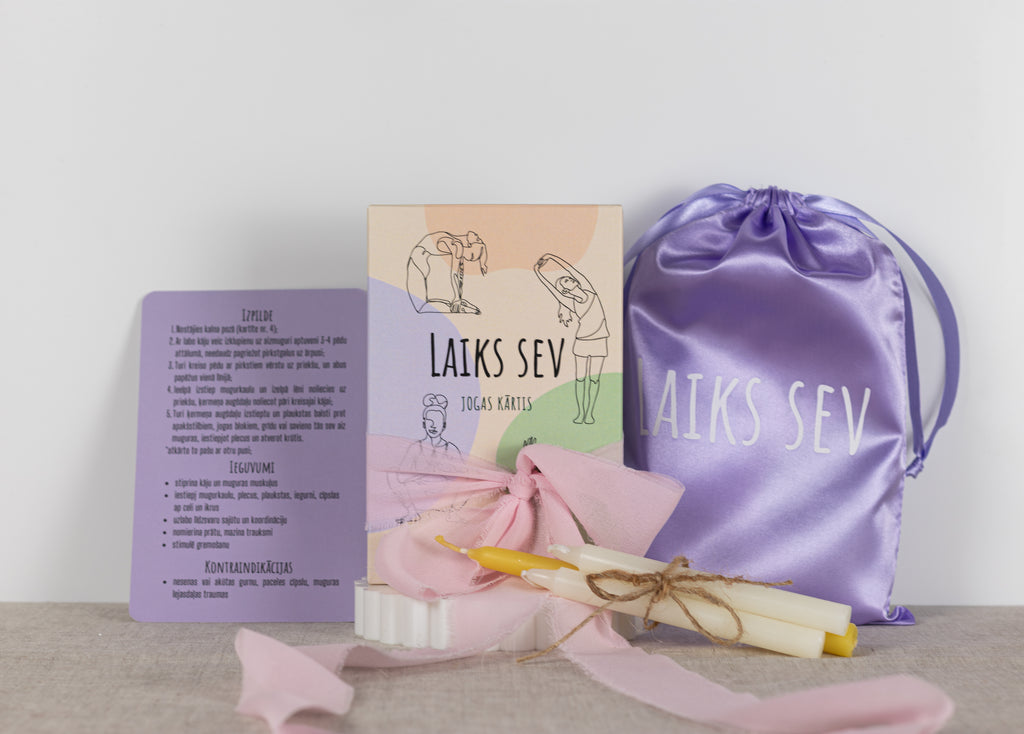 A gift set featuring a lavender satin drawstring bag with 'LAIKS SEV' printed in white, a lavender instruction card, a pink ribboned box with a card, and two yellow-tipped candles tied with twine, arranged on a neutral surface against a light background.