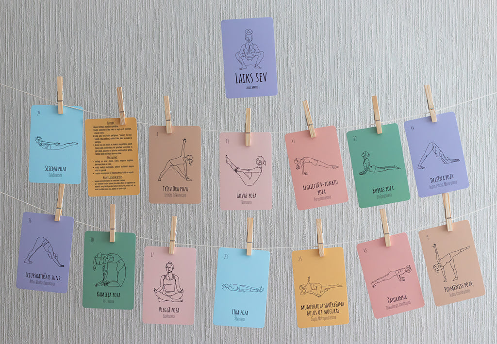 A set of colorful yoga pose cards clipped to strings against a light gray textured wall. The cards feature minimalist line art and text for various yoga poses, including Svan Mula, Tadasana, and Lotus Pose.
