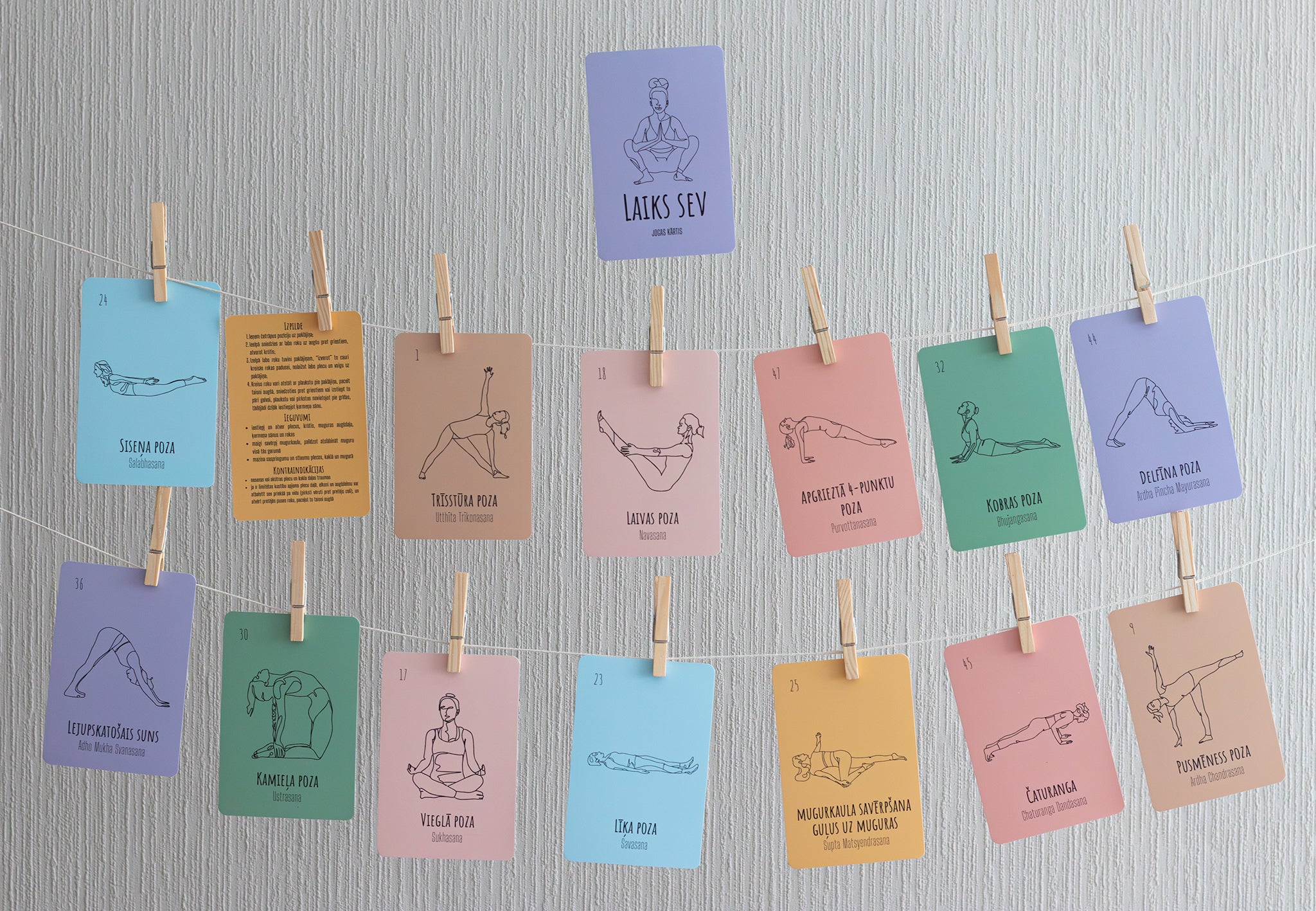 A set of colorful yoga pose cards clipped to strings against a light gray textured wall. The cards feature minimalist line art and text for various yoga poses, including Svan Mula, Tadasana, and Lotus Pose.