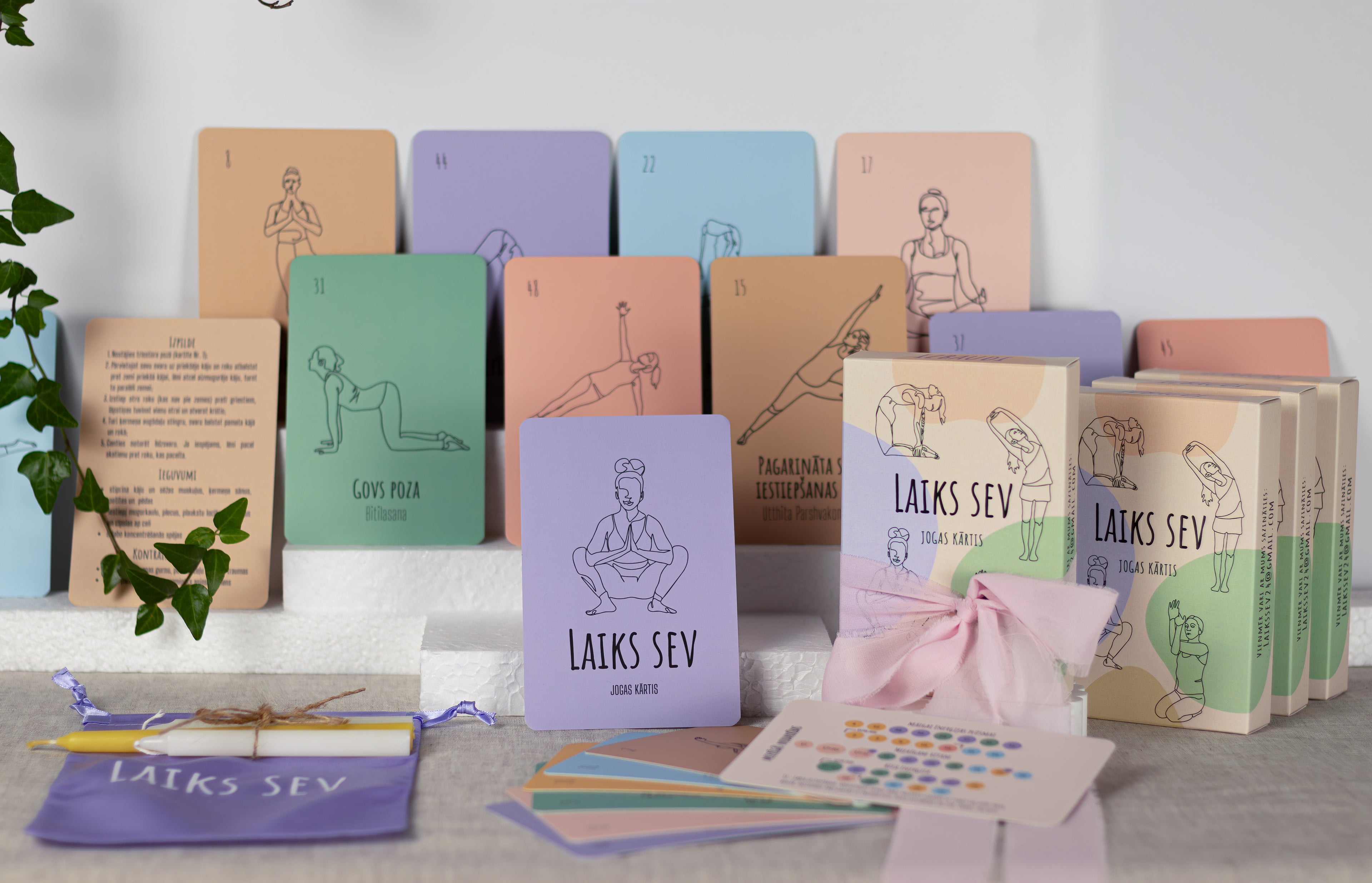 A set of pastel-colored yoga and meditation cards, books, and a small pouch labeled 'LAIKS SEV' arranged on a light surface against a white wall, with a plant leaf visible on the left.
