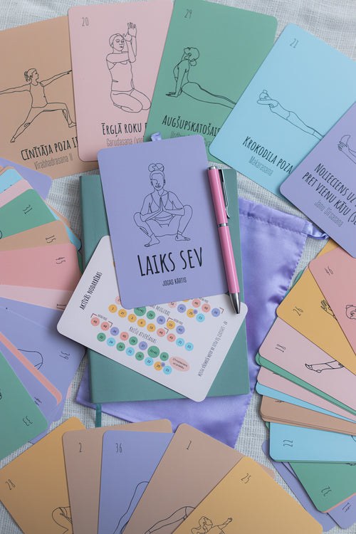 A set of pastel yoga and meditation cards arranged in a flat lay, featuring illustrated figures in various poses and Latvian text such as 'LAIKS SEV' and 'SVEKA TUKU'. A pink pen and a small lavender pouch are placed on top of the cards.