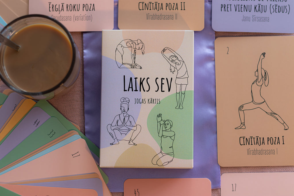 A yoga card deck titled 'LAIKS SEV' (Your Own Yoga Cards) displayed on a purple surface, surrounded by cards showing different yoga poses. A glass of coffee is visible in the top left corner.